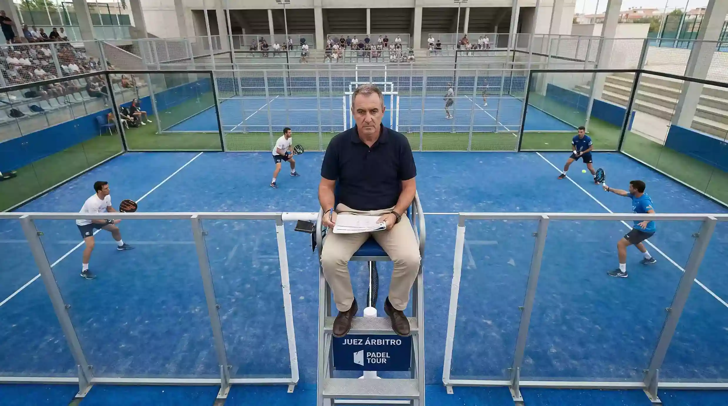 Árbitro de pádel en su silla elevada supervisando un partido profesional en una pista de cristal
